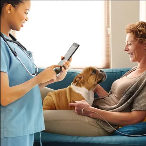 A doctor in scrubs wearing a stethoscope reads from a tablet device while talking to a patient laying on a comfortable couch petting a dog while receiving an IV infusion.